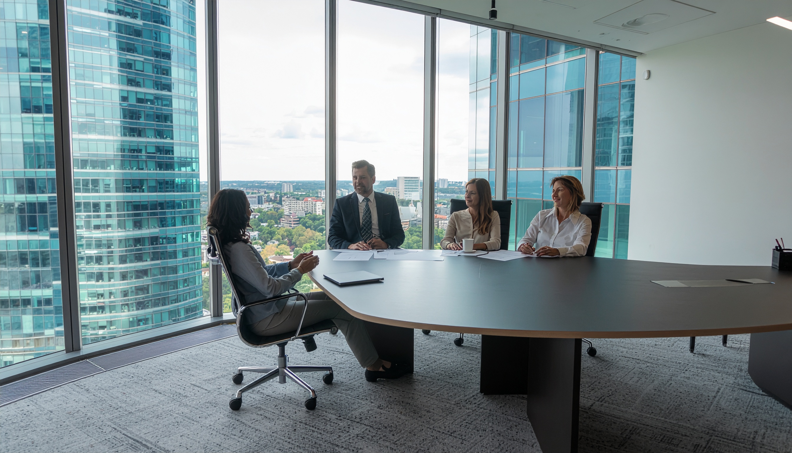 Sala de reunião moderna em arranha-céu, com quatro pessoas em trajes formais sentadas ao redor de uma mesa oval. Janelas amplas oferecem vista panorâmica da cidade, criando um ambiente iluminado e profissional. O foco está na interação do grupo, sugerindo uma discussão de negócios produtiva. A composição destaca linhas verticais dos edifícios ao fundo.